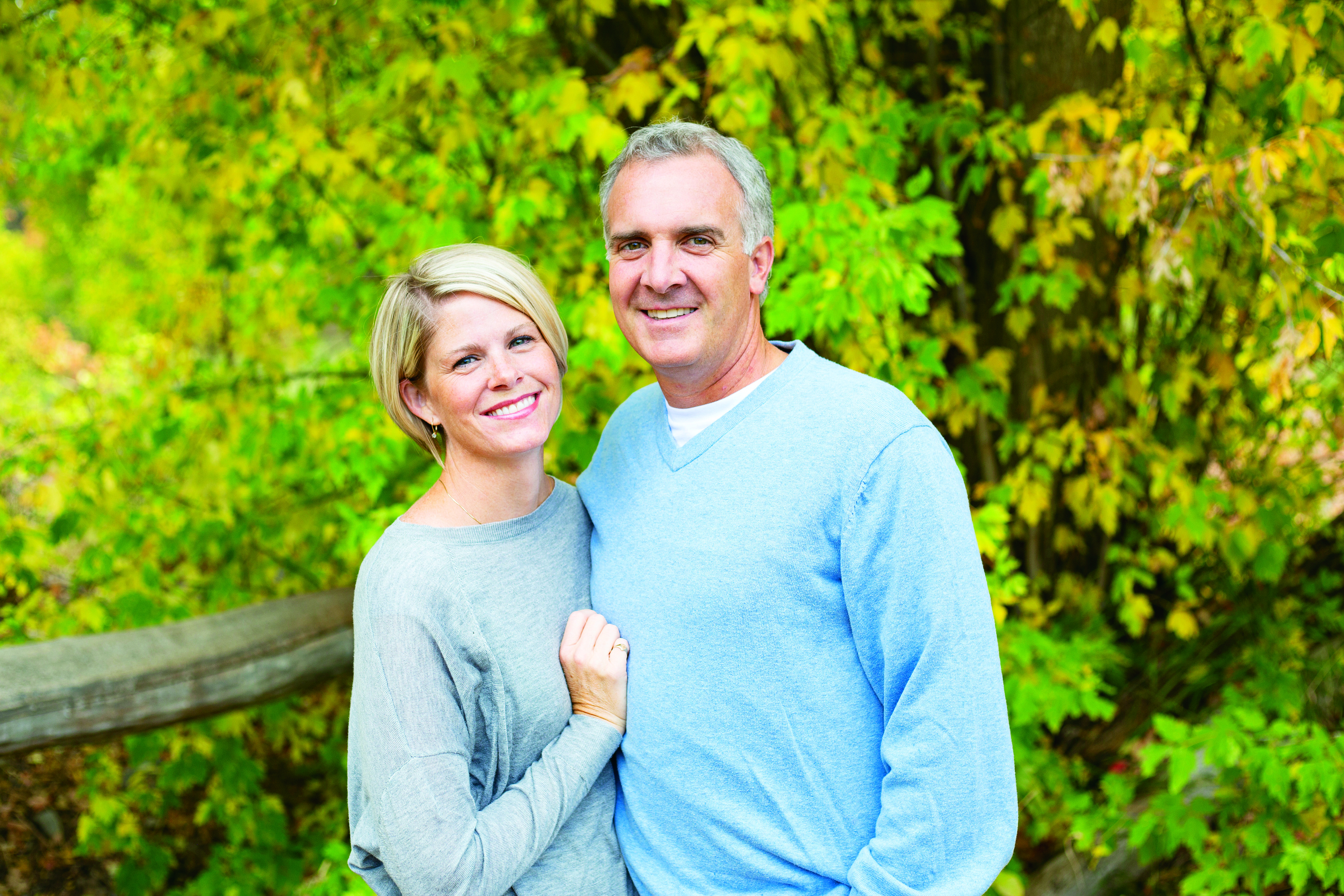 A smiling couple is standing in front of bright green bushes.