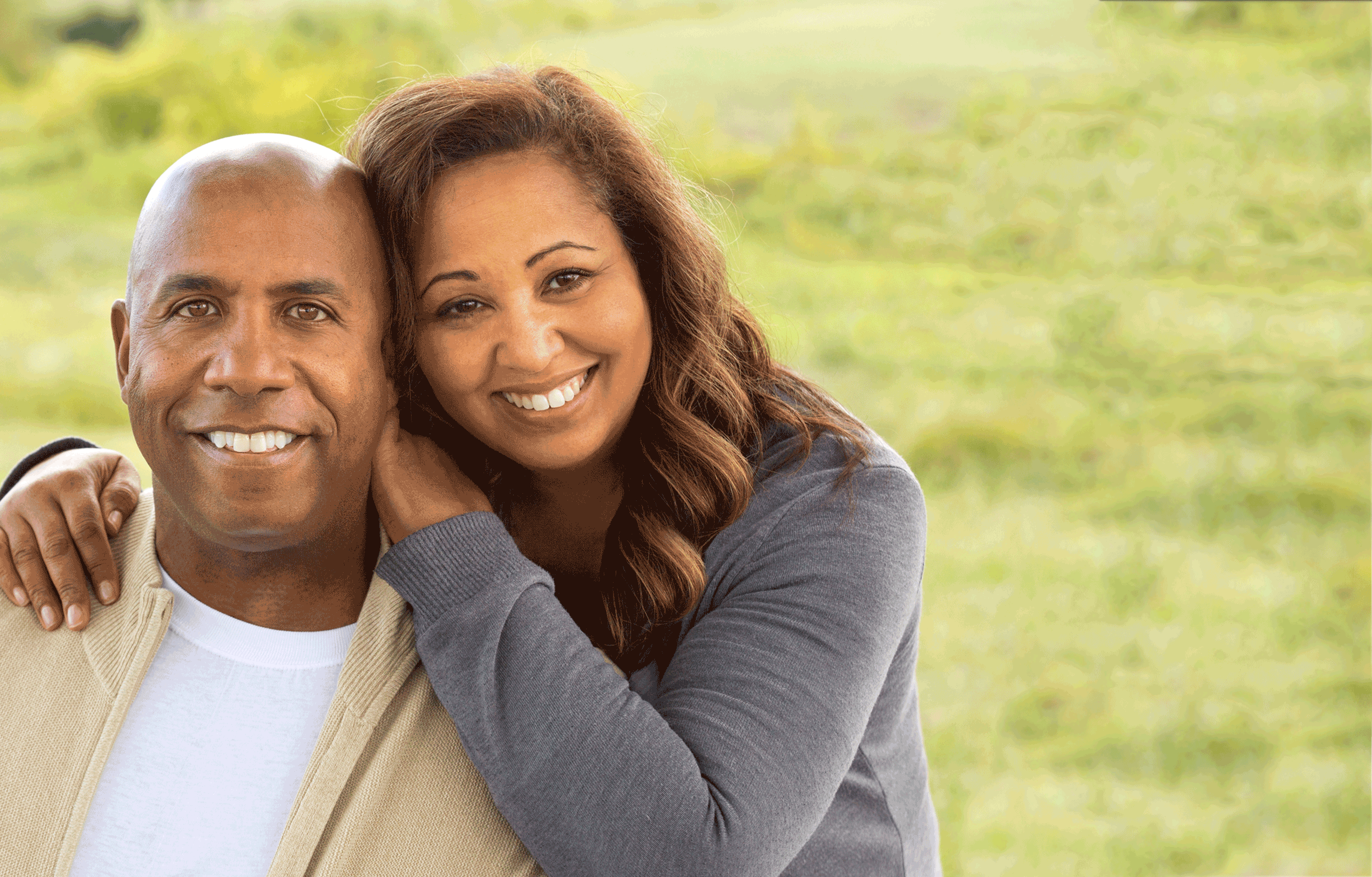 A smiling couple is in front of a grassy field.