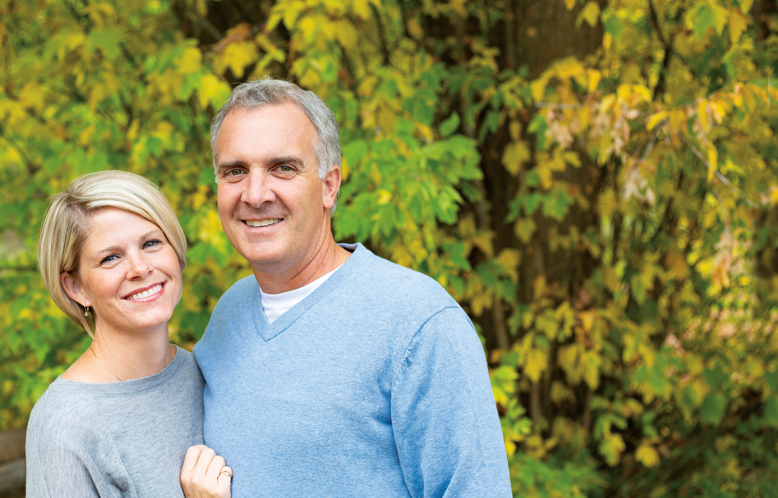 A smiling couple is standing in front of green and yellow trees.