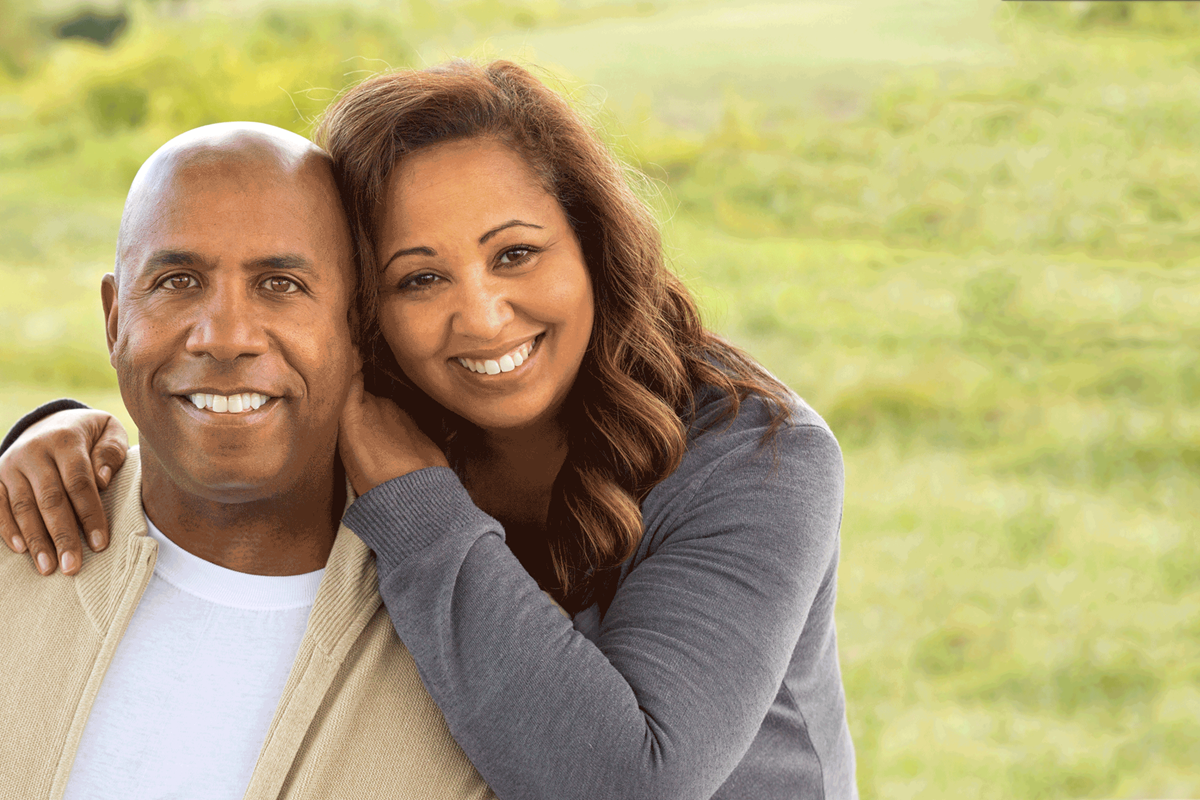 A smiling couple is in front of a grassy field.