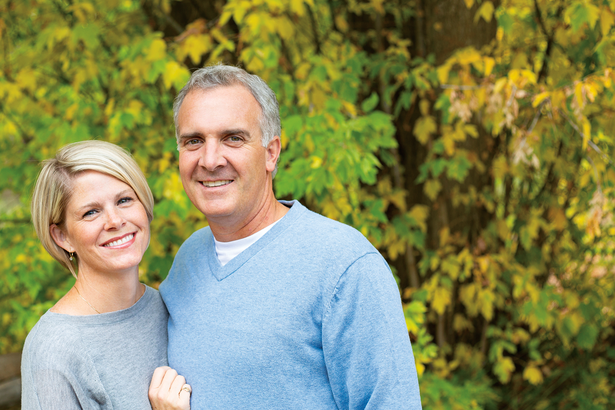 A smiling couple is standing in front of green and yellow trees.